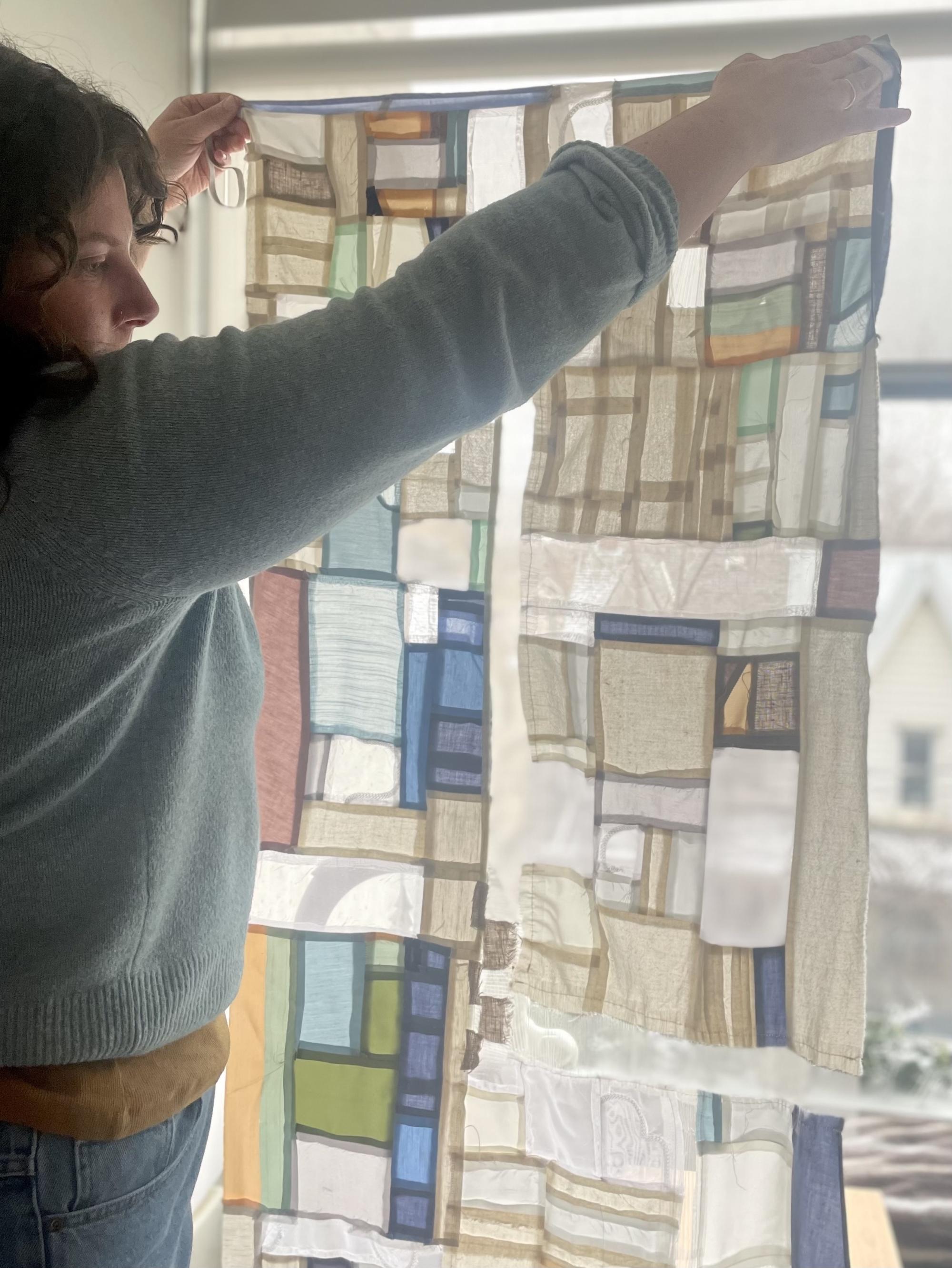 A young woman stands in front of a window, holding up a quilt-like patchwork artwork. The light from the window illuminates the colourful patches of the artwork, highlighting its intricate designs.