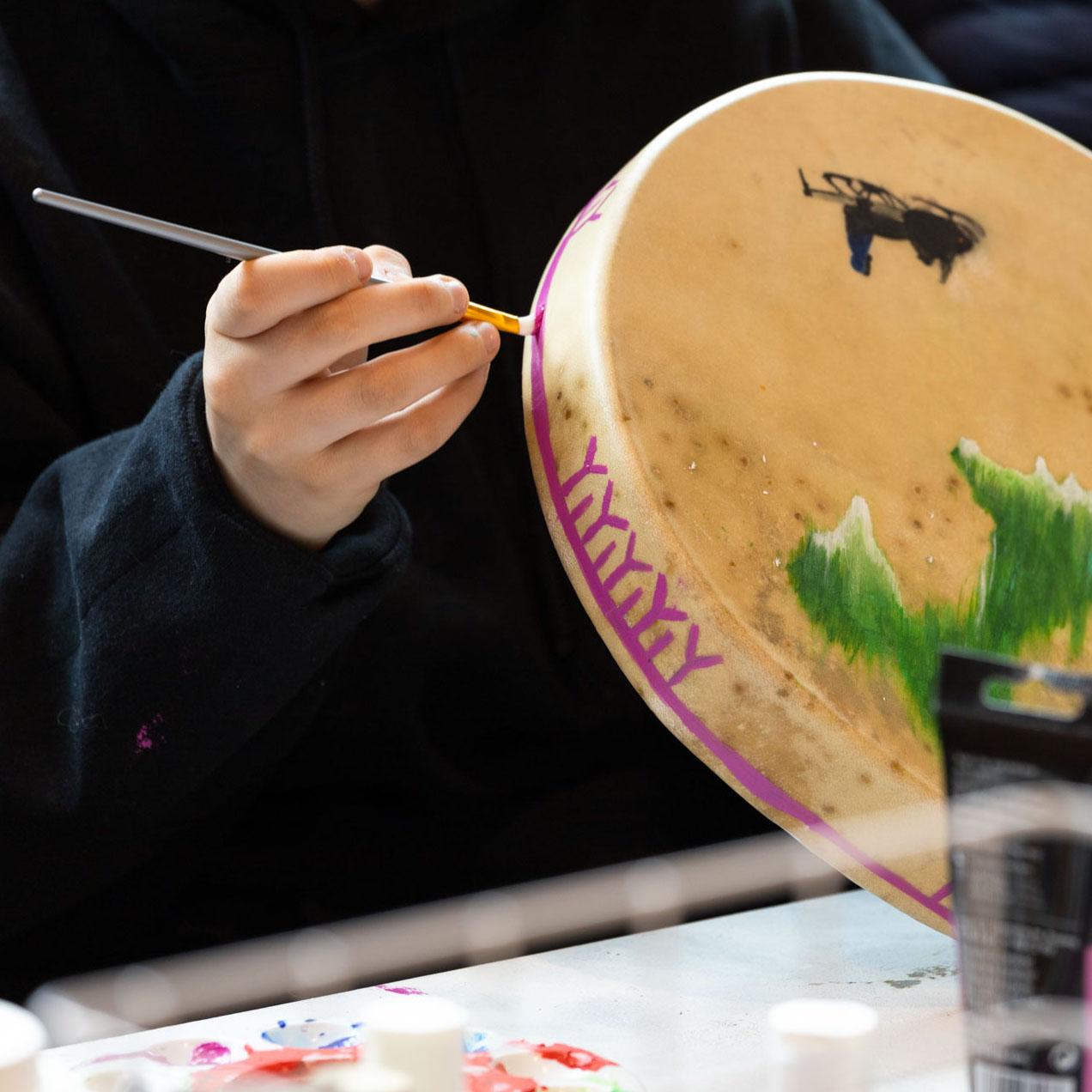 Raw hide drum side being painted with pink Inuit designs showing the front painted northern-lights above snowmobile