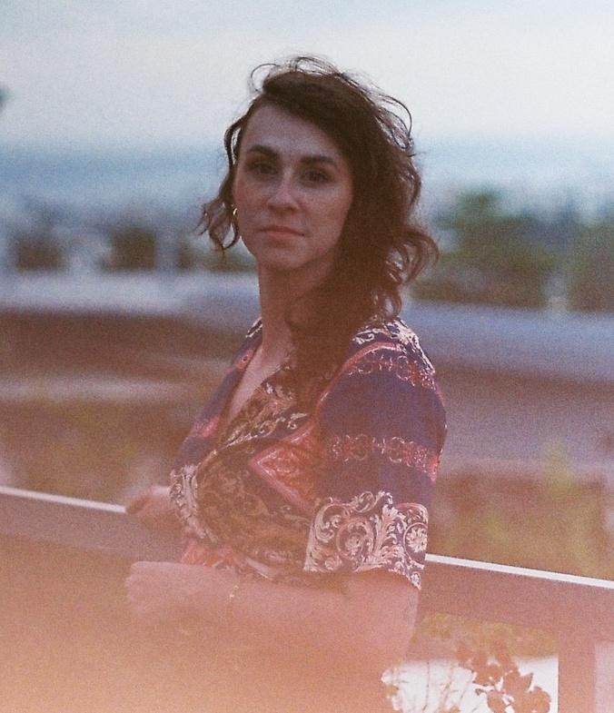 A caucasian woman with brown hair faces to the left and looks at the camera. She is wearing a multicoloured shirt. The photo is slightly distressed. 