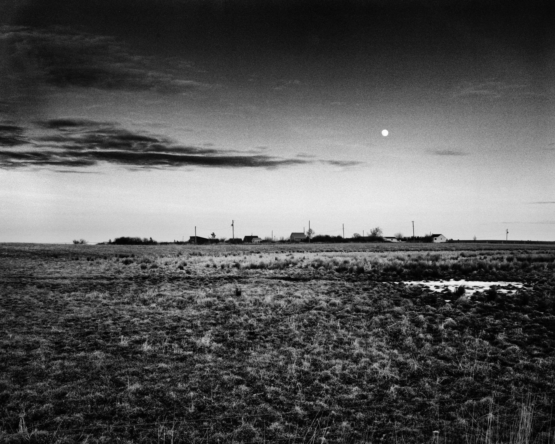 A black and white landscape photograph of a street of houses in the middle of a large grass field. The houses occupy the centre of the photograph in the distance while overhead the moon appears in the sky as the sun begins to set 