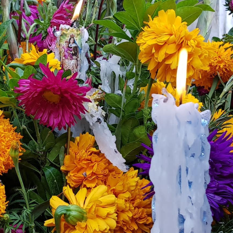 photograph of a white wax candle with a bright orange flame, surrounded by brightly coloured pink, orange, and yellow flowers