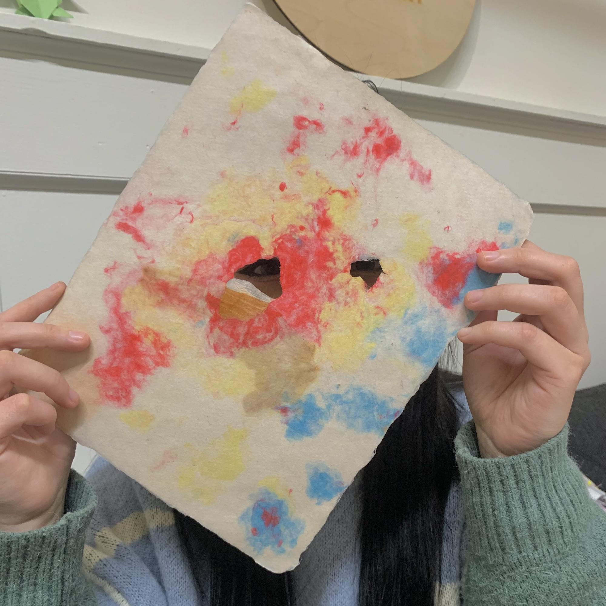 a person holds a piece of handmade paper in front of their face. Their eyes are visible peeking out from two holes in the paper. The paper has red, yellow and blue splotches in it. 