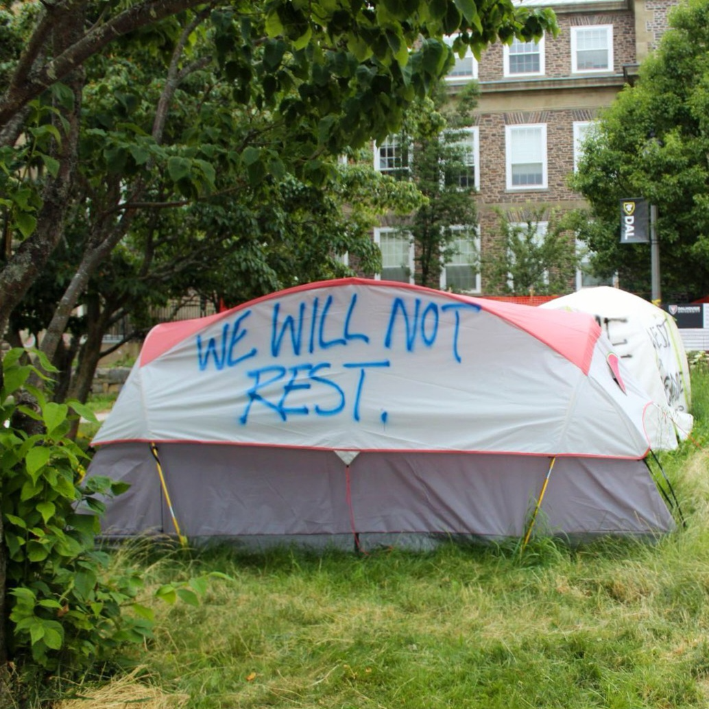 photograph of a gray and red tent nestled in a grassy area with trees and shrubs around it, in front of a brick building. "We will not rest" is spray painted in blue capital letters along the side of the tent