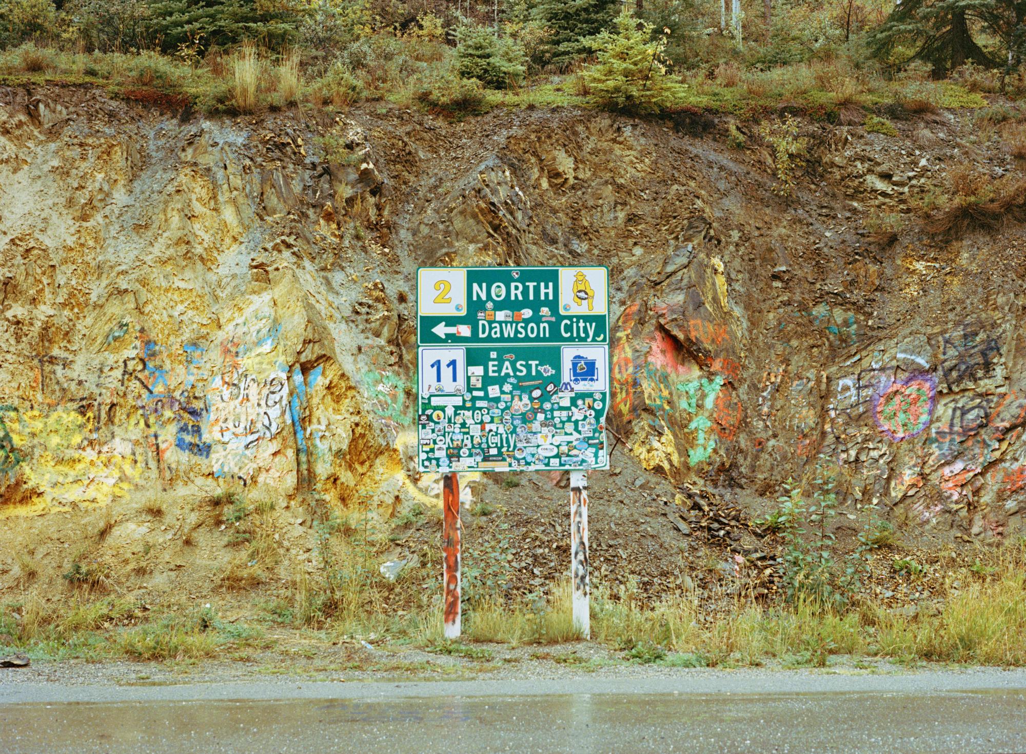 Flat view of a road sign of Dawson City, covered in stickers, and placed in front of a rock cliff face covered in graffiti.