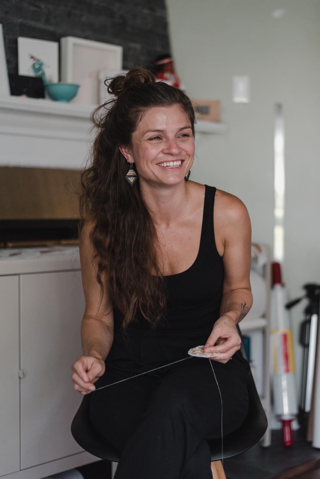 photo of Carrie sitting on a stool with a needle and thread in her hand as she beads, she is smiling looking to the right