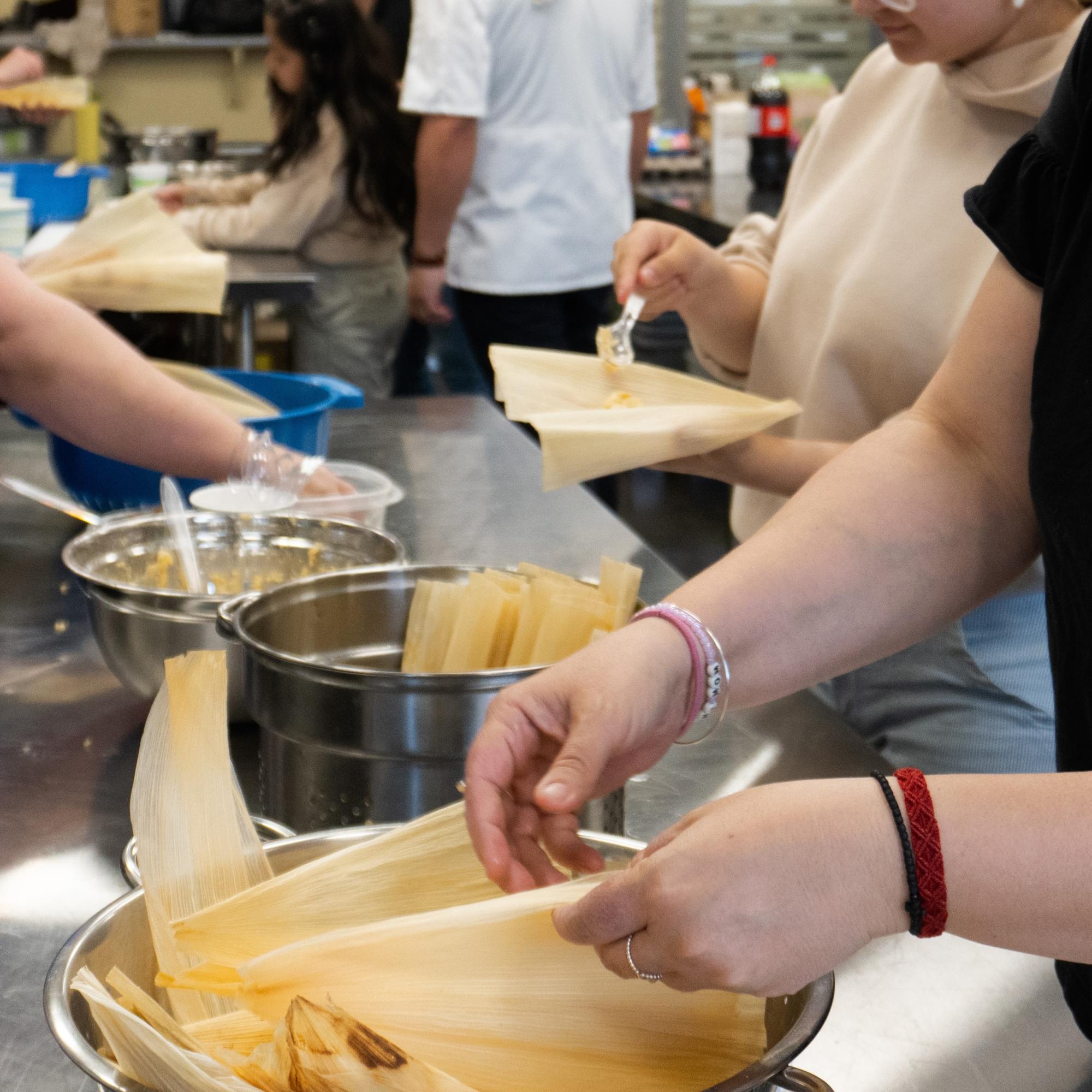 photograph of peoples hands grabbing corn husk wraps and filling them with the mixture to make tamales