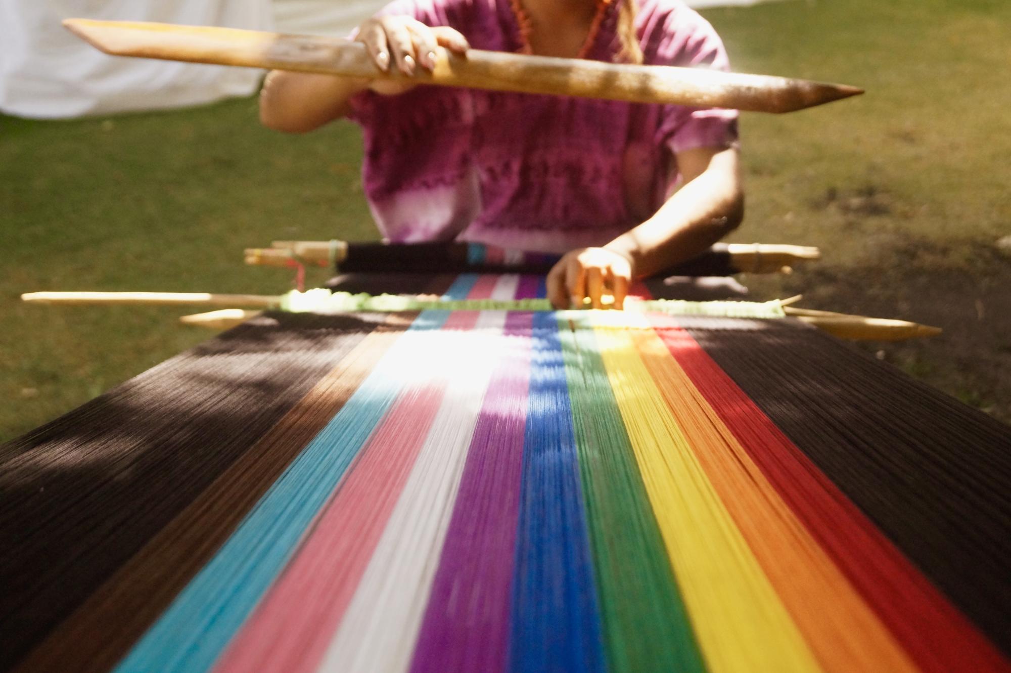 photograph of a person weaving on a large loom. The strands of textiles are in the colors of the pride flag