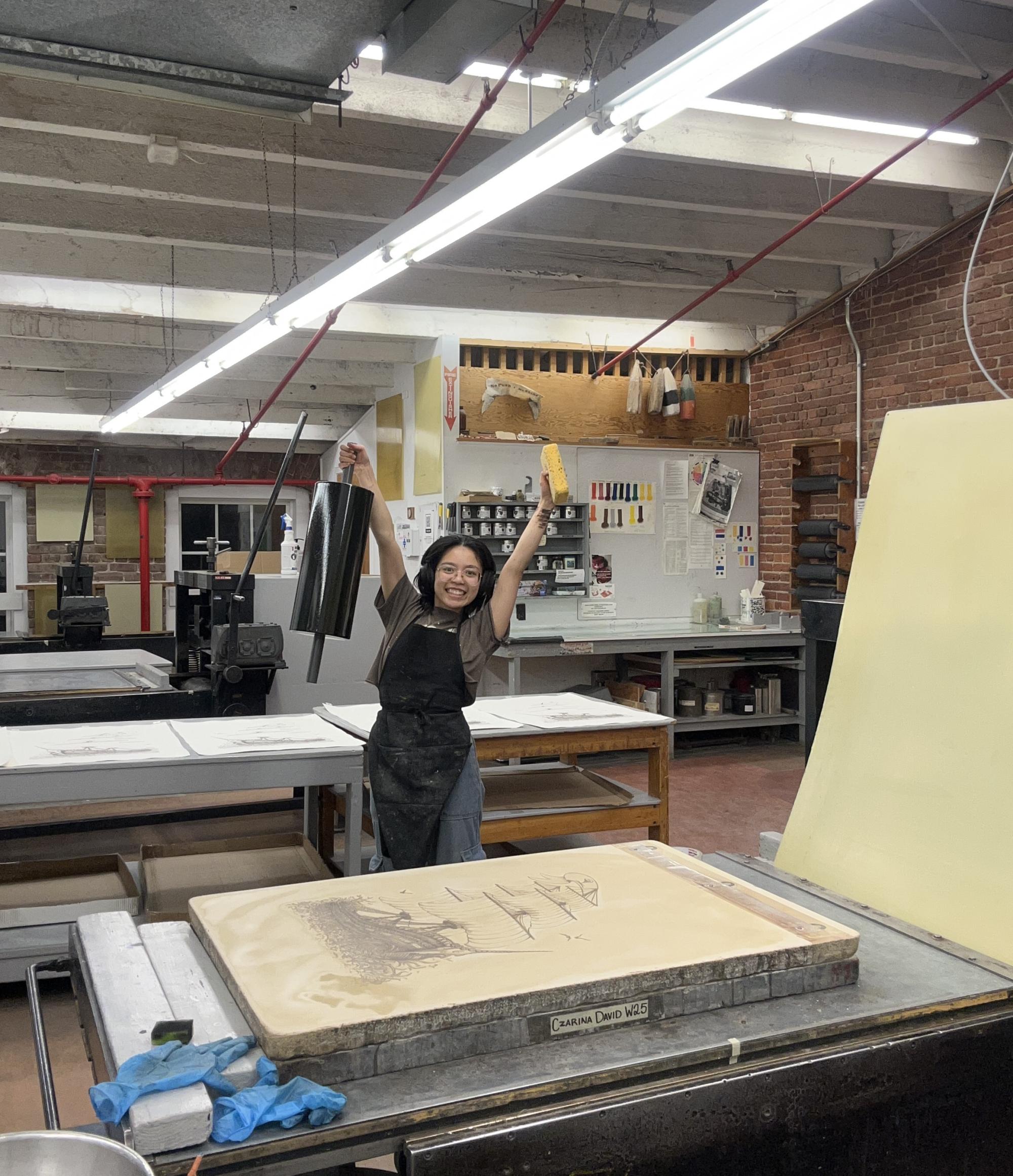 Czarina holding a big litho roller and sponge. Holding her hands in the air, next to a litho stone and press.