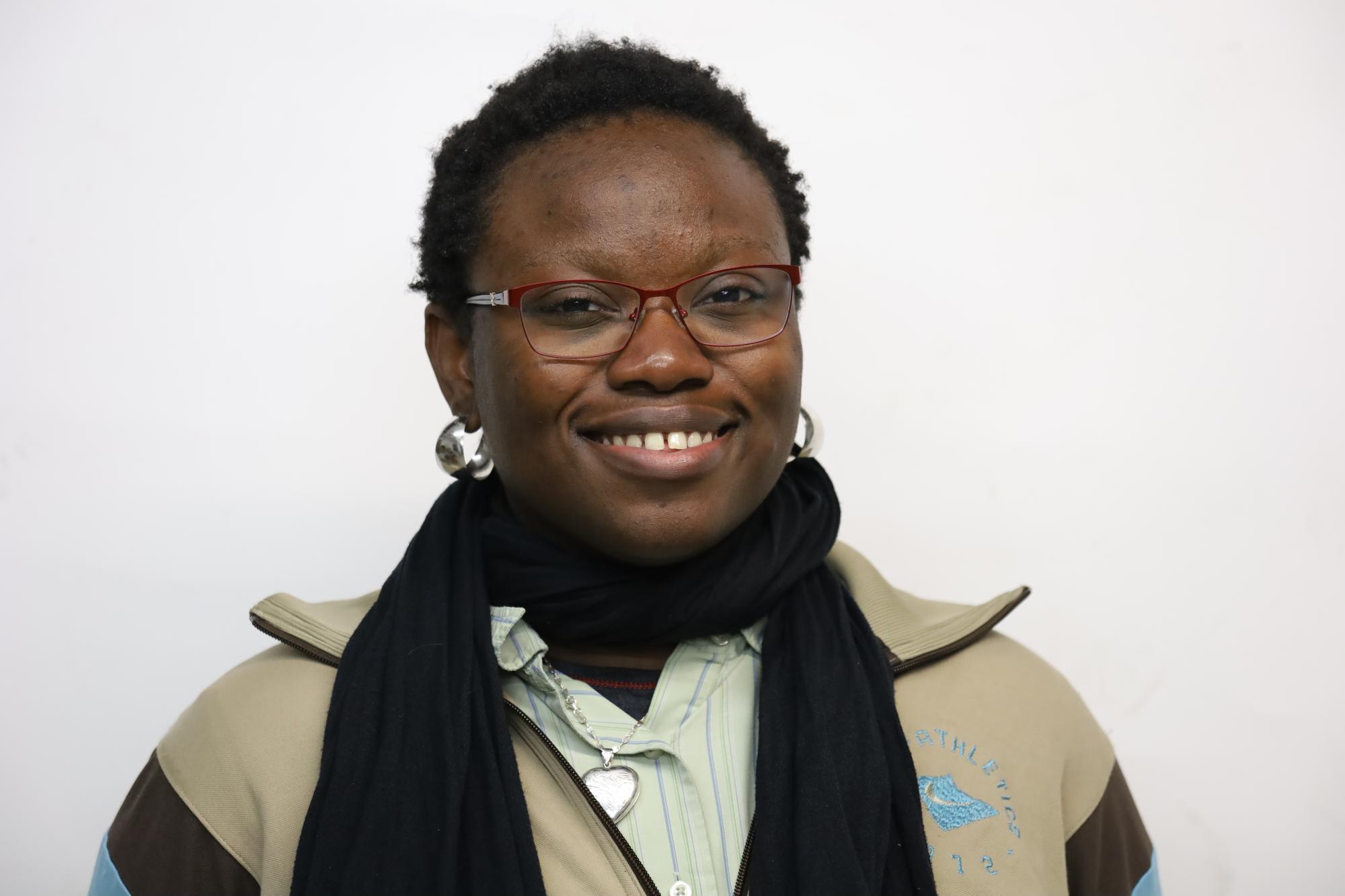 Busola stands in the centre of photograph smiling at camera. She has black hair, brown eyes and is wearing thin red glasses, a black scarf and a striped shirt under a beige and brown colour block track jacket 