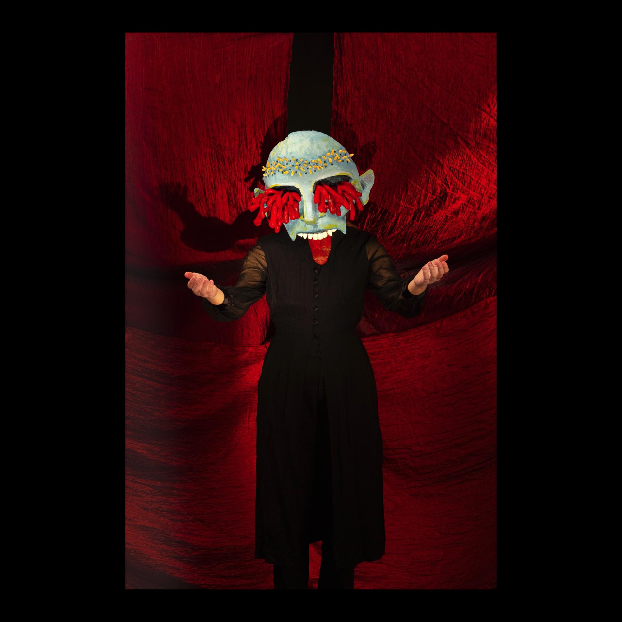 Death Mask, person wearing a blue skinned mask with red guts coming out of the eyes and a maggot crown, standing in front of a red curtain