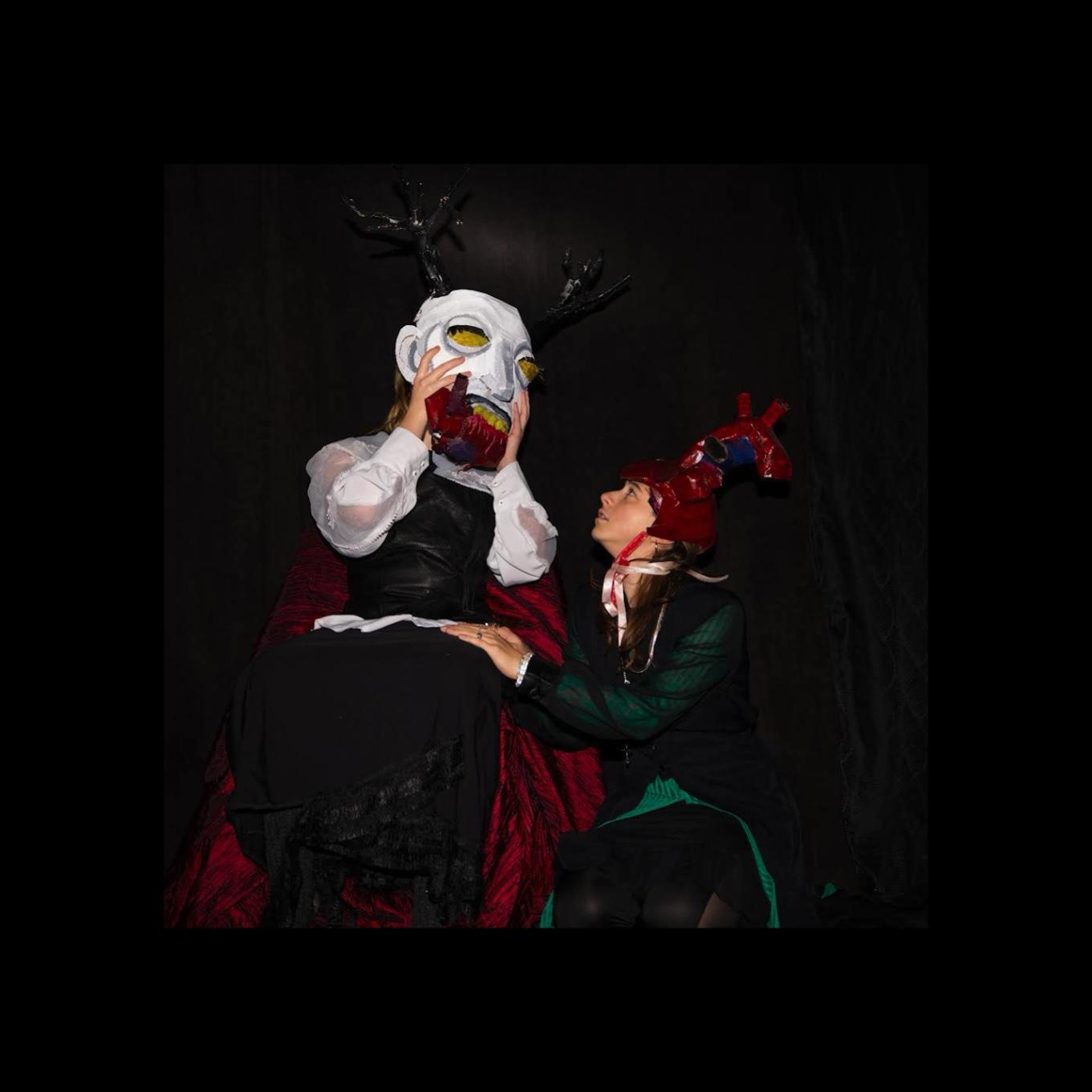 Sinnerman (7/7), a antlered, white face being with a red heart in their mouth stare at a knelt person with a anatomical heart mask on their head.
