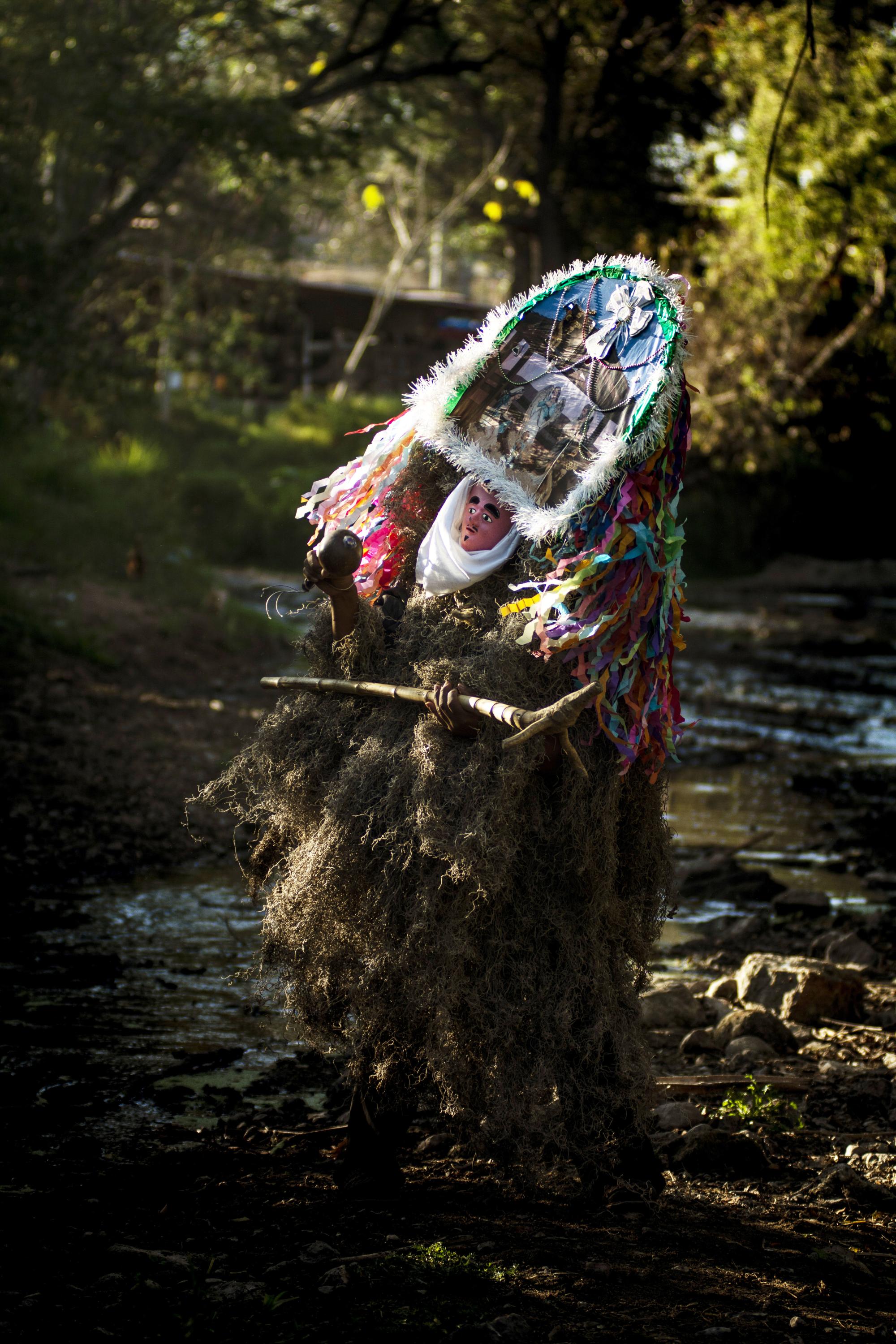 A costumed figure dances in the woods wearing a mask that resembles an altar and robe covered in plant materials, holding a pick axe like traditional tool.