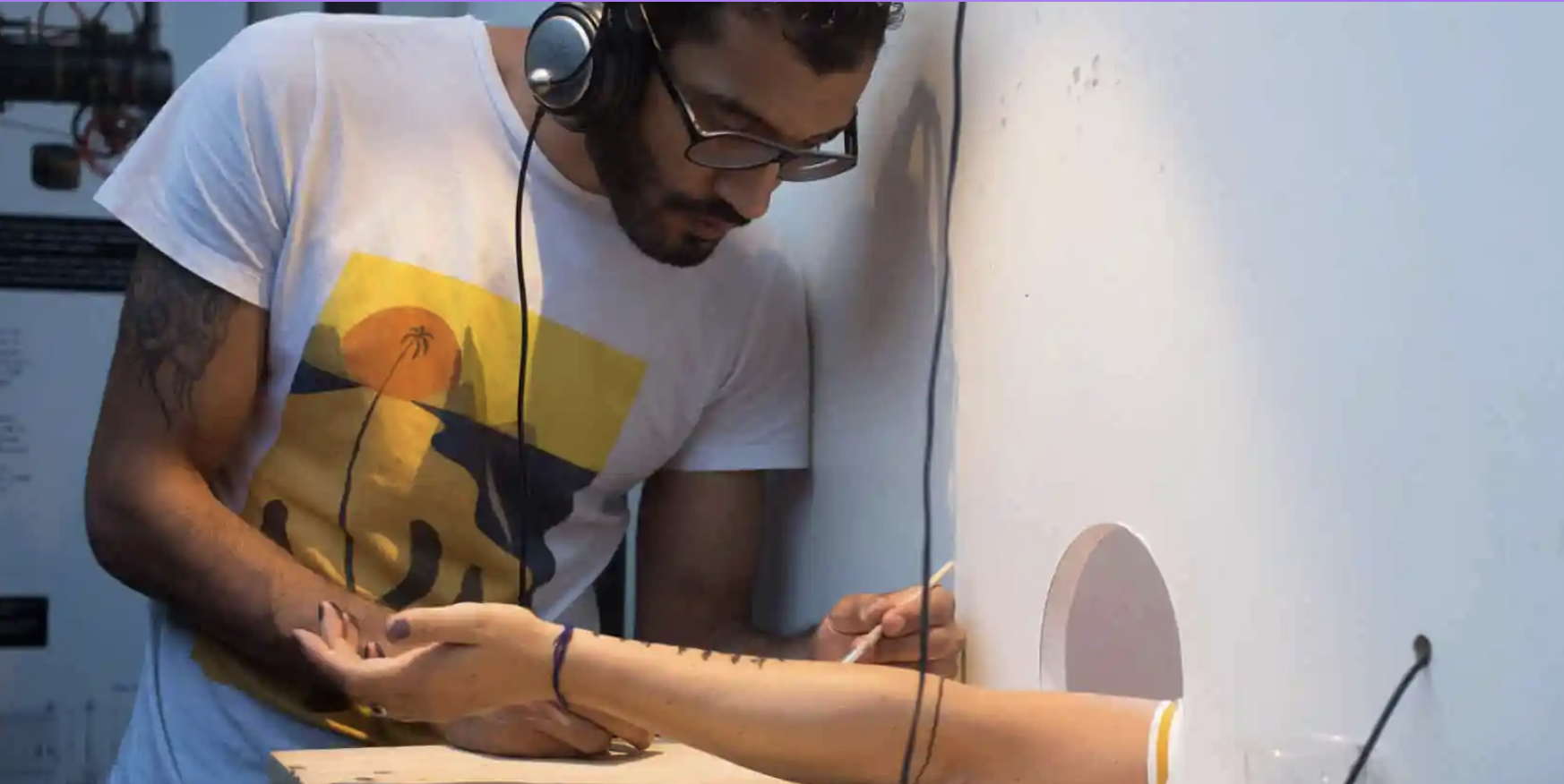 An arm protrudes from a hole in the wall hand upturned. A person of colour wearing glasses and headphones is drawing on it.