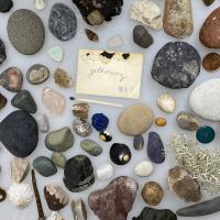 A collection of small stones, shells, seaweed, buttons, matches, a penny, an old nail, sea glass, around the word gathering.