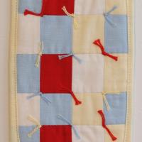 a small, rectangular quilt the size of a doctor's prescription pad. Featuring colours from a prescription label - red, yellow, white and blue. The quilt consists of small, colourful squares and has cotton thread ties binding the quilt together.