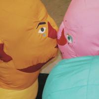 Two large, brightly coloured soft sculpture heads are arranged to kiss each other. The bright orange head, on the right of the frame, has a blue heart tattoo on their face. The pink head is angled slightly away from the viewer, and has eyelashes and beauty marks.