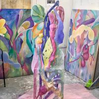 Brightly coloured sculptural work stands vertically in the middle of a studio space, with colourful paintings rest against walls. The sculpture features green, purple, pink, orange and yellow shapes. The paintings appear to be interpretations of the colourful sculpture.