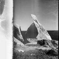 Two large boulders in front of the ocean, that look as if they were split from one boulder. The image is in black and white film and there are fluid looking distortions on the surface of the film. On the left side of the film the image is white with grey drip marks.