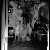 A monochromatic black and white photograph of a monstrous, humanoid creature crouched in the corner of a dimly lit furnace room. 