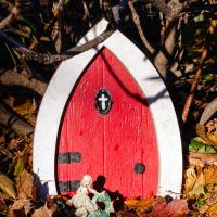 An oval-shaped, wooden, red door with thick white trim sits on a carpet of leaves beneath bare branches of shrubbery. Black metal hinges, a black doorknob, and a flat black oval door knocker with a white painted cross adorn the door. A small figurine is situated in front.