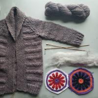 Wool, granny squares, knitting needles, and yarn laid next to a hand knit purple cardigan, comprised of the materials. 