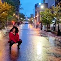 A person wearing black leggings and high heels with a bright red blazer and long black curly hair crouches in the middle of the street, looking at the camera. The sky is a dark blue and the street glistens with rain. The Halifax Citadel Hill clock tower can be seen in the background at the top of the street. 