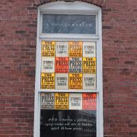 Neon orange letterpress prints hang in the window of a brick building. Prints read "Letterpress is a tool of colonization.”
