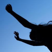 A person reaching their arms ups. The image is rotated so the horizon line is vertical. They are backlit with a blue sky.