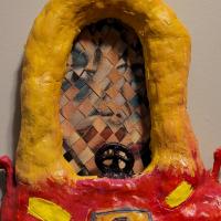 A woven painting of a human face, framed within a papier-mâché and paper clay toy car sculpture painted in acrylic. 