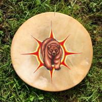 [ID: "Square photo of a natural coloured rawhide drum, with a painting of a brown bear with a red and white coloured design behind the bear - similar to a sun. The drum is placed on bright green grass."]