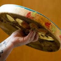 A tattooed arm holds out a rawhide drum, stamped with a strawberry design on its outer edge. The rawhide lacing on the back of the drum is visible.