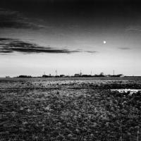 A black and white landscape photograph of a street of houses in the middle of a large grass field. The houses occupy the centre of the photograph in the distance while overhead the moon appears in the sky as the sun begins to set 