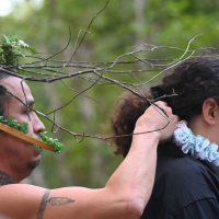 photograph of two performers, Richard "Krow Dog" Taylor, costumed in a basket with leaves and branches and Kayli Raye Marr wearing a lace ruff