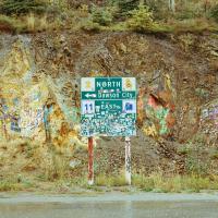 Flat view of a road sign of Dawson City, covered in stickers, and placed in front of a rock cliff face covered in graffiti.