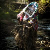 A costumed figure dances in the woods wearing a mask that resembles an altar and robe covered in plant materials, holding a pick axe like traditional tool.