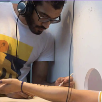 An arm protrudes from a hole in the wall hand upturned. A person of colour wearing glasses and headphones is drawing on it.