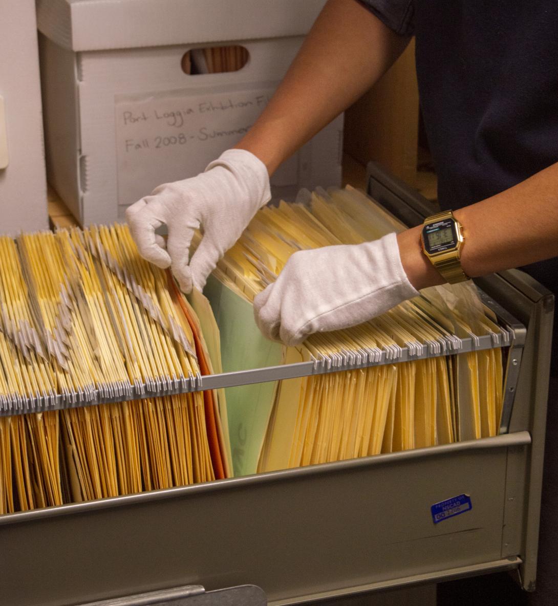 a dark skinned person wearing white gloves searches through a filing cabinet of yellow manilla folders