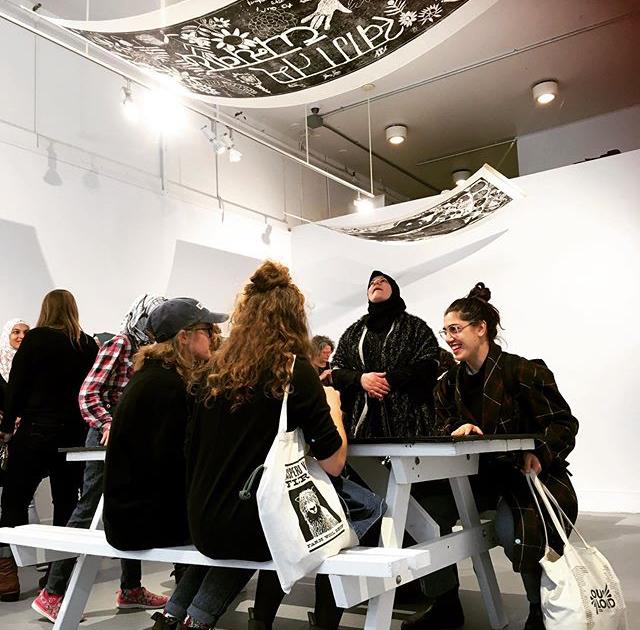 a group of people sitting at a picnic table in the gallery under woodcut prints hung from the ceiling. 